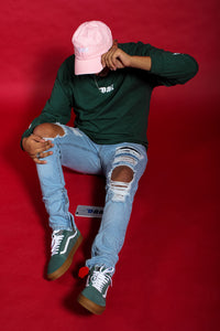 Person wearing a green sweatshirt, pink cap, and ripped jeans sitting on a red background
